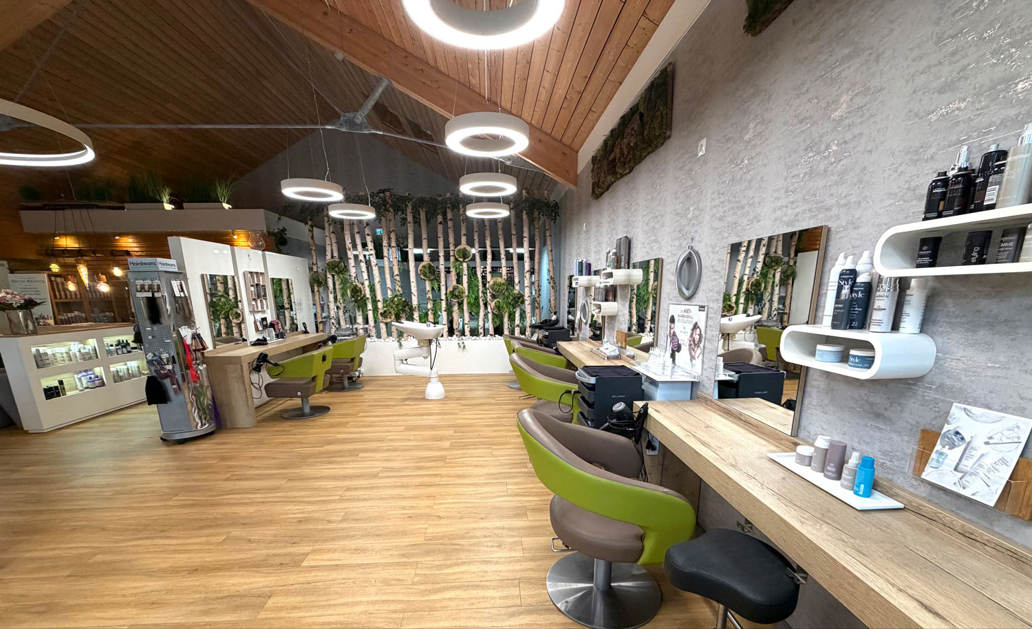 Blick durch den Salon: Bedienplätze mit grünen Stühlen, Holzboden und Ringleuchten